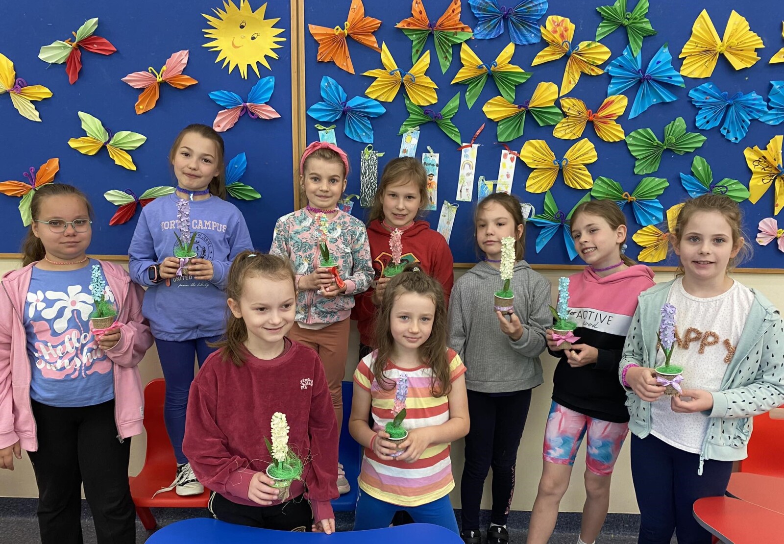 Dziewięć dziewczynek stoi przed tablicą z motylami i pokazuje zrobione przez siebie hiacynty z papieru w kolorowych doniczkach.
