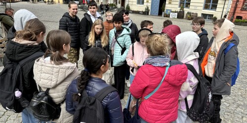 Uczniowie wraz z przewodnikiem i opiekunem stoją na rynku głównym w Lublinie. W tle zabytkowe, malownicze kamienice.