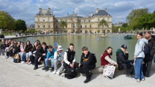 Dziewczynki i chłopcy siedzą na murku przy fontannie przed Pałacem Luksemburskim.