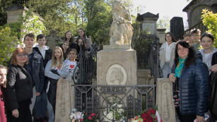 Grupa dziewczynek i chłopców wraz z opiekunami stoi przy pomniku nagrobnym Fryderyka Chopina znajdującym się na cmentarzu Père-Lachaise.