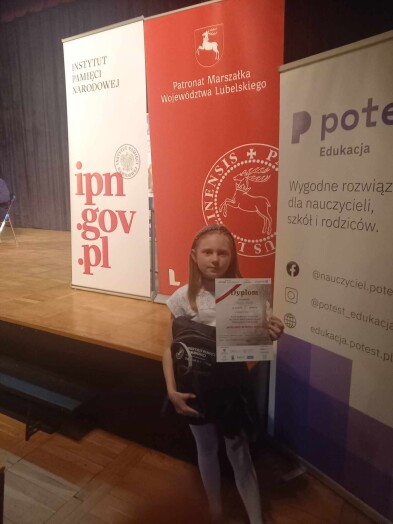 Sukces w Konkursie Poetyckim „Niezłomni w moich oczach” 2 – uczennica trzyma dyplom na tle banerów